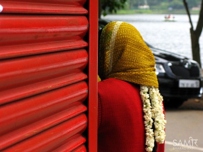 Back of a woman wearing a  scarf and a string of flowers in her hair, leaning on shop shutters